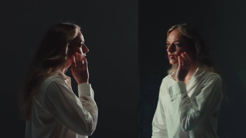 A troubled blonde woman in a white shirt slowly touches her face as she confronts her reflection in a dark, moody mirror