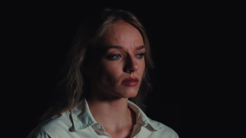 Close-up portrait of a beautiful, sad young woman with striking blue eyes looking thoughtfully off-camera against a dark background