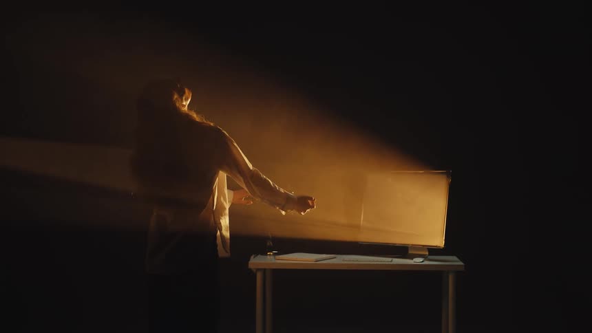 A woman in a dark, hazy room slowly flexes her advanced cybernetic prosthetic hand in the warm glow of a screen