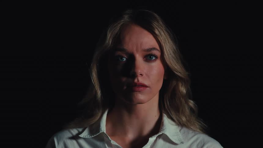 A dramatic close-up portrait of a serious blonde woman with blue eyes staring intently at the camera in a dark room