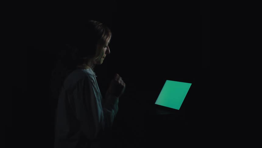 A young woman sits in a dark room, her face illuminated by the glow of her green-screen laptop as she works
