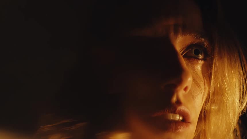 An intense close-up captures a woman's terrified face illuminated by a flickering, warm light in slow motion