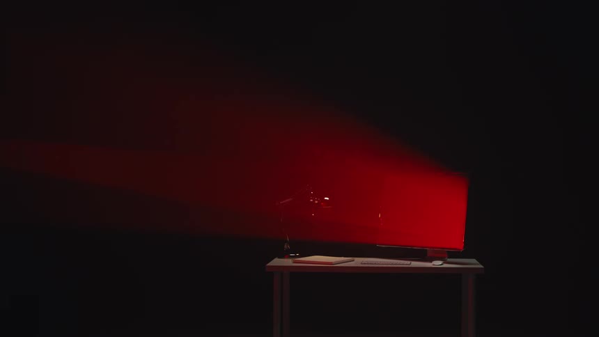 An empty desk with a computer, lamp, and notepad is illuminated by a dramatic red light in a dark room