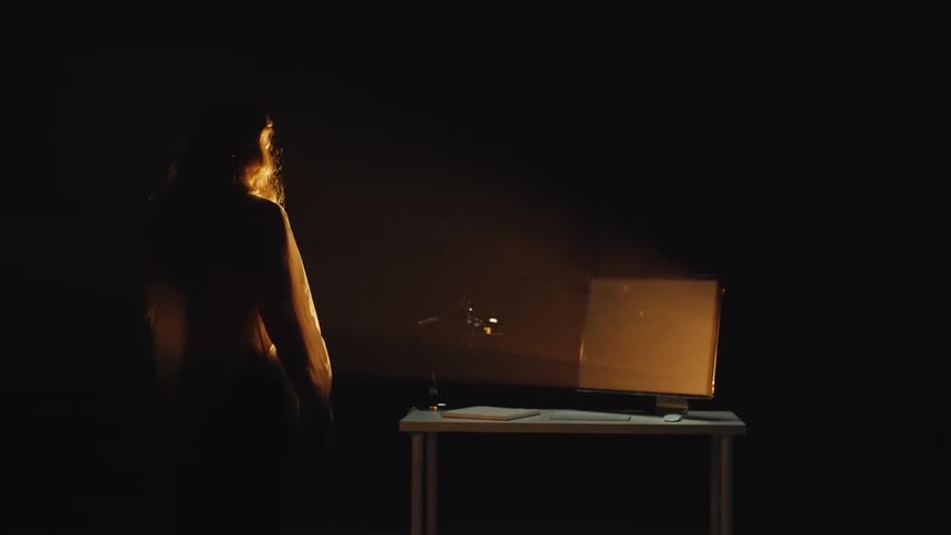 In a dark room, a woman is drawn towards the glowing light of a computer screen