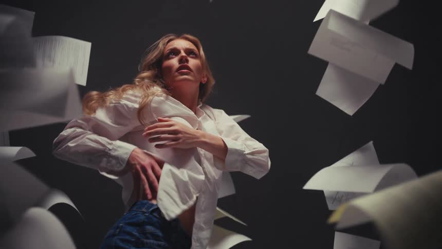 A woman performs an expressive, frantic dance in slow motion, surrounded by a chaotic whirlwind of floating papers