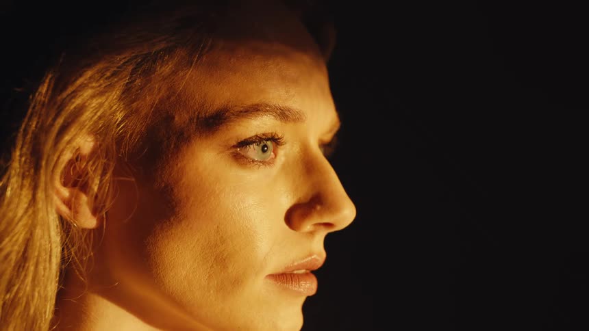 A woman's face in profile, illuminated by warm light against a black background, slowly opens her eyes and looks up with awe