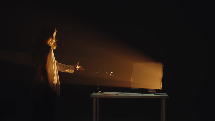 A woman in a dark room makes slow, deliberate gestures towards a computer screen illuminated by a single warm spotlight