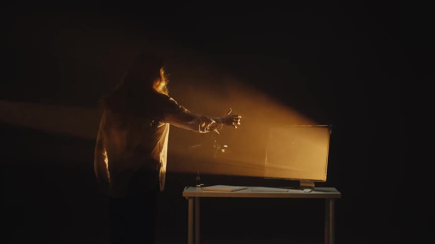 A woman in a dark, smoky room slowly approaches an old television and reaches her hand out towards its glowing, empty screen