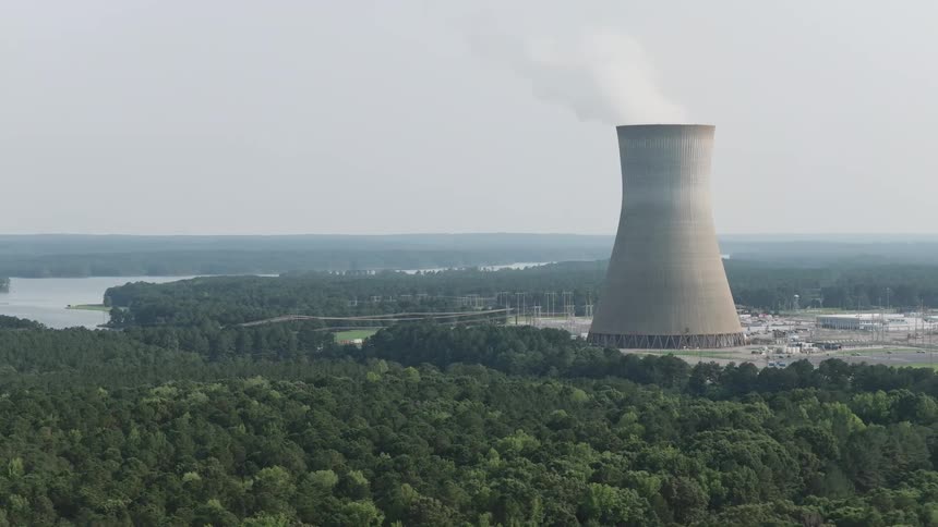 Wide shot of a nuclear power plant's cooling tower with steam rising above a dense forest and a nearby lake