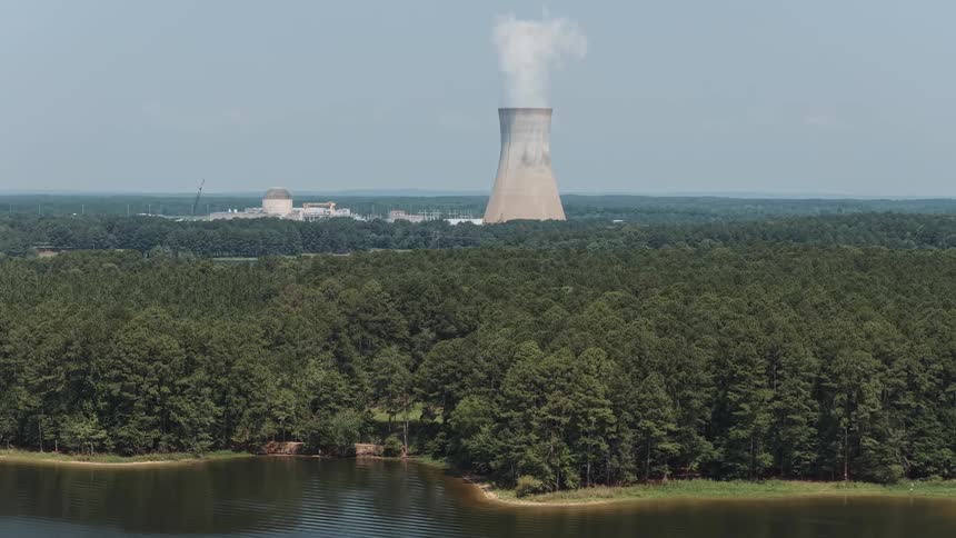 A nuclear power plant's cooling tower releases steam over a dense forest landscape bordering a calm lake