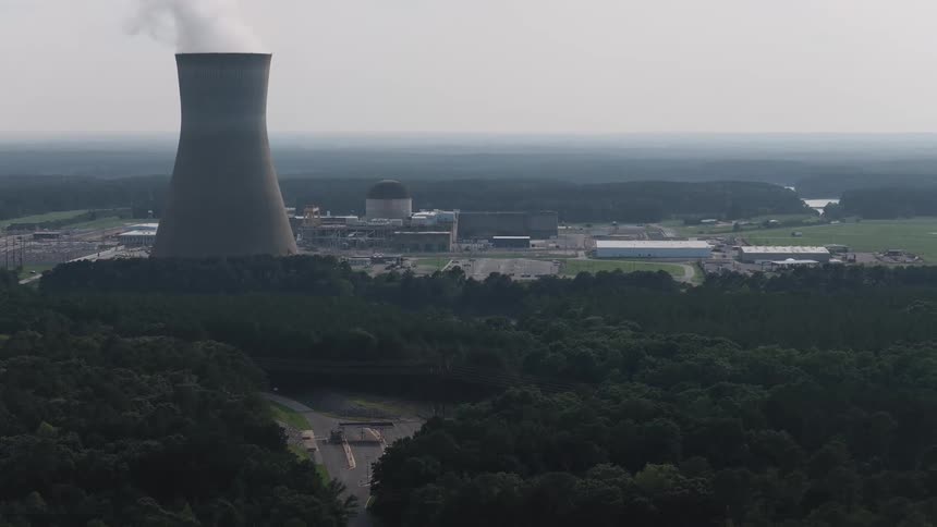 Slowly pulling back aerial shot of a nuclear power plant with a steaming cooling tower surrounded by a lush green forest
