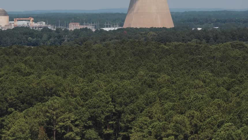 Aerial view rises from a tranquil lake shore over a vast forest to reveal a nuclear power plant with steam coming from its cooling tower
