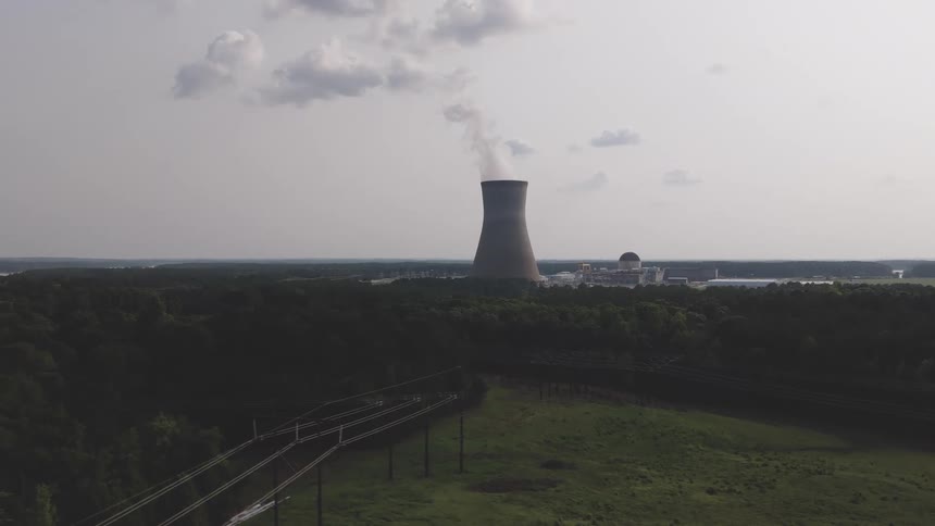 An aerial shot slowly approaches a nuclear power plant and its cooling tower in a rural, wooded area