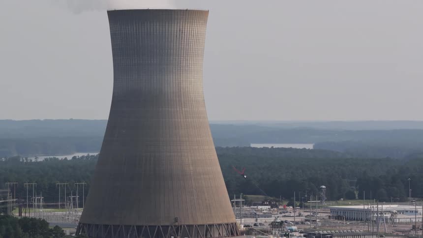 A slow upward pan reveals a massive nuclear power plant cooling tower emitting a steady plume of steam into the overcast sky, with the industrial facility and surrounding forest below