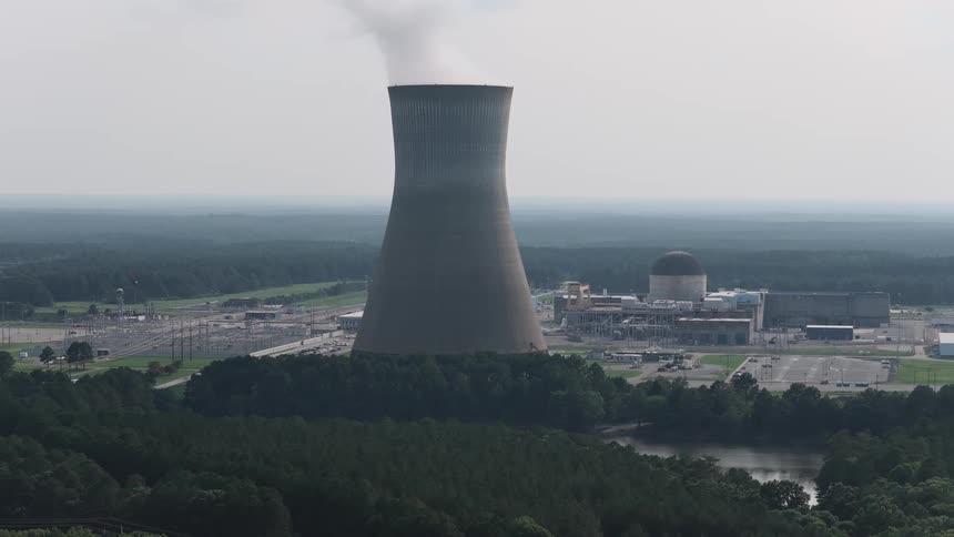 A slow aerial pull-back shot of a nuclear power plant with steam rising from the cooling tower in a rural, forested landscape