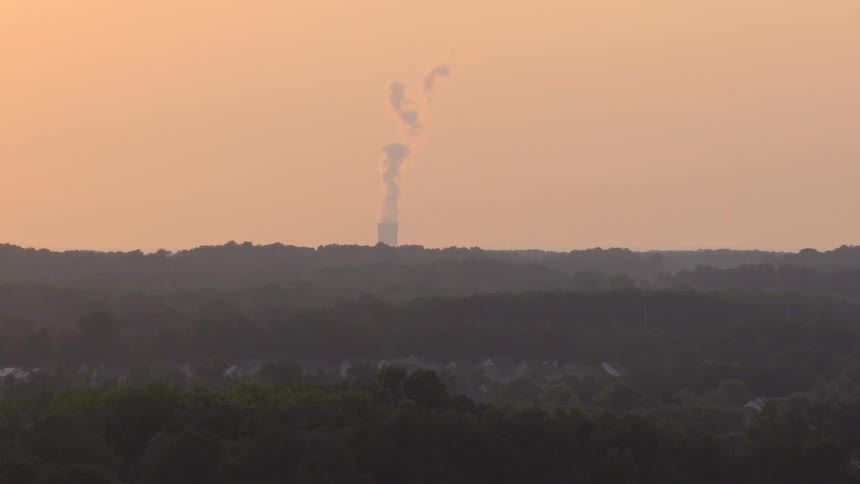 A slow zoom on a power plant's cooling tower emitting steam over a vast, hazy forest at sunset