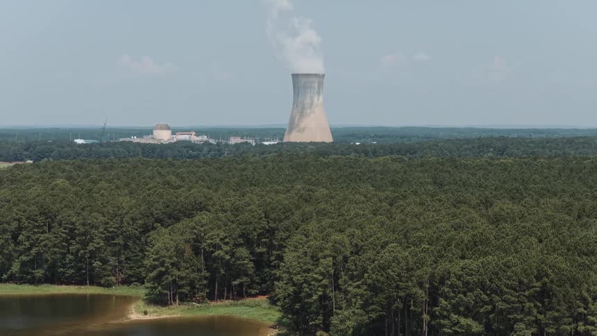 An aerial shot pulls back from a nuclear power plant emitting steam over a dense forest to reveal a large, calm lake in the foreground