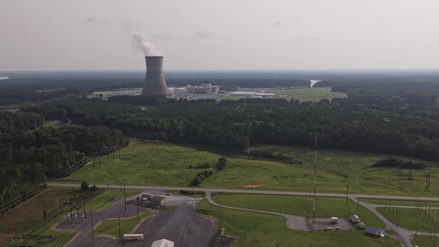 A slow aerial push-in towards a nuclear power plant, with a cooling tower emitting steam above a vast green, rural landscape