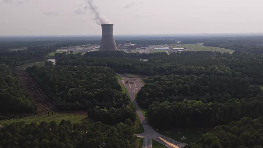 An aerial shot slowly reveals a nuclear power plant with steam coming from its cooling tower, surrounded by a dense forest