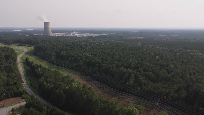 A slow aerial pullback reveals a nuclear power station with a steaming cooling tower surrounded by a vast rural forest and power lines