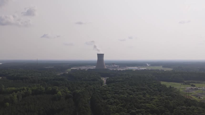Slow aerial reveal of a nuclear power plant's cooling tower emerging from behind a dense forest landscape