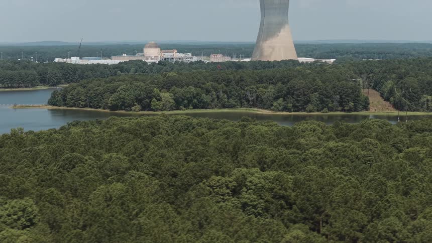 Aerial reveal of a nuclear power plant and cooling tower with steam rising over a vast green forest and lake