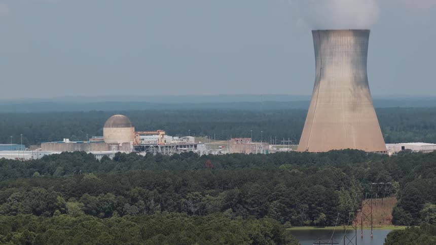 A wide shot of a nuclear power plant with a prominent cooling tower emitting steam, surrounded by a dense green forest and a lake