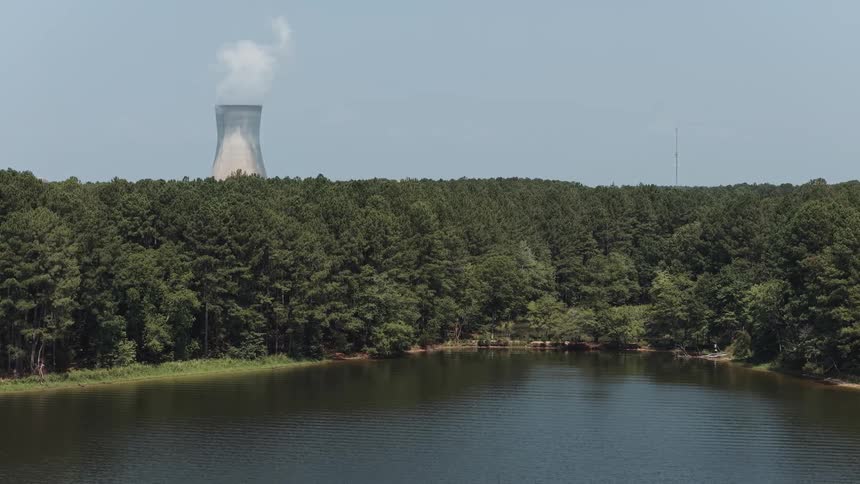 A slow aerial shot moves across a serene lake and dense forest, revealing a nuclear power plant cooling tower emitting steam in the distance