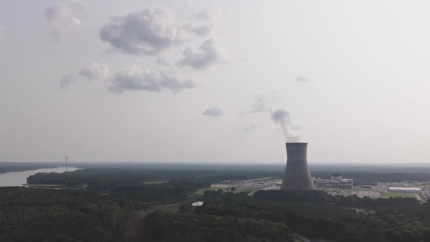 A wide aerial view of a nuclear power plant with a cooling tower releasing steam into the hazy sky, set amidst a dense forest next to a river