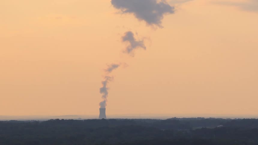 Distant view of a nuclear power plant cooling tower emitting a plume of steam against a hazy orange sky at dusk