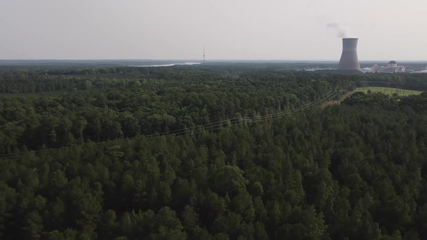 A slow, wide aerial view reveals a nuclear power plant's cooling tower emitting steam above a vast, dense forest