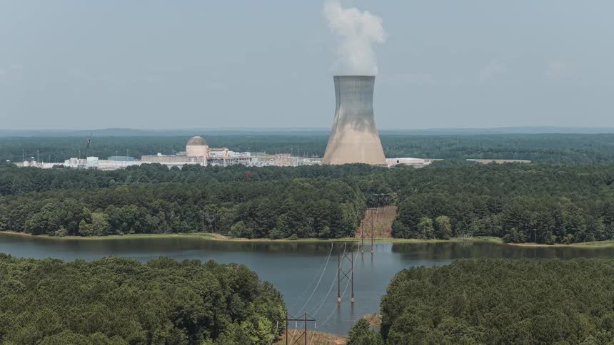 An elevated view of a nuclear power plant with a large cooling tower emitting steam, situated on the edge of a lake and surrounded by dense green forest
