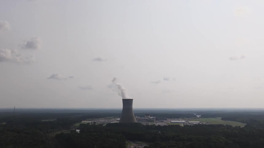 A slow aerial tilt-down reveals a nuclear power plant's cooling tower emitting steam into a hazy sky, surrounded by a dense forest