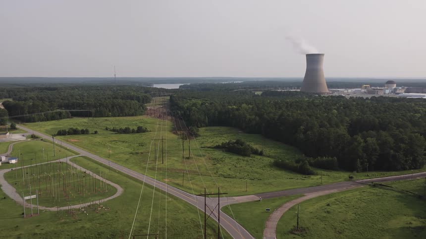 Cinematic aerial footage pulls back over power lines and rural roads to reveal a nuclear power plant with its cooling tower emitting steam