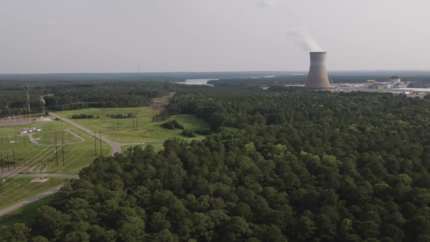 Slow aerial flight over a dense green forest landscape towards a nuclear power plant with steam rising from a cooling tower