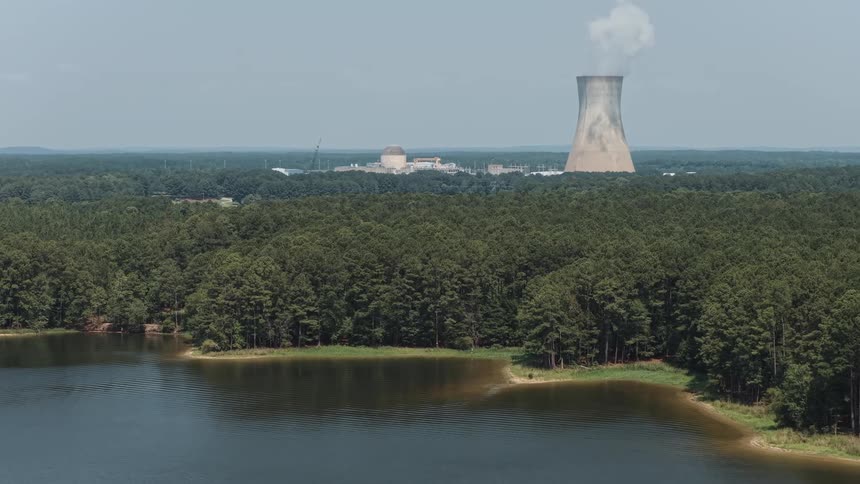 A nuclear power plant with a smoking cooling tower stands in the distance behind a dense forest and a tranquil lake