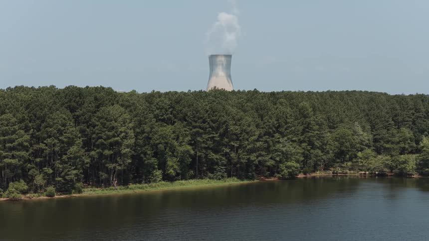A nuclear power plant cooling tower steams in the distance behind a lush forest and a calm lake on a clear day