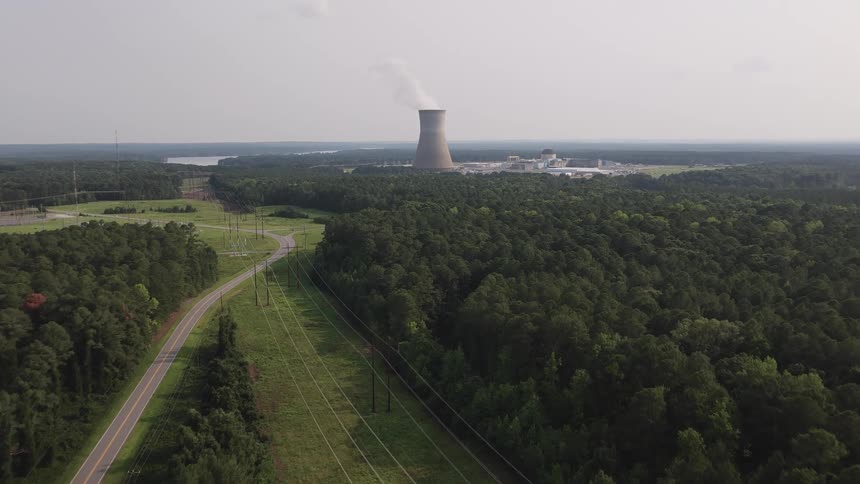 An aerial drone shot slowly advances over a vast, dense forest and power lines towards a distant nuclear power plant
