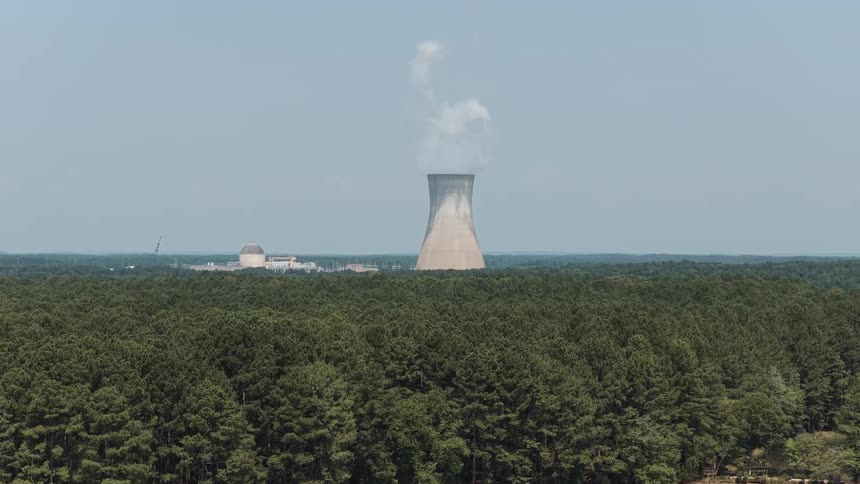 An aerial shot slowly pulls back from a nuclear power plant's cooling tower over a dense forest to reveal a large lake