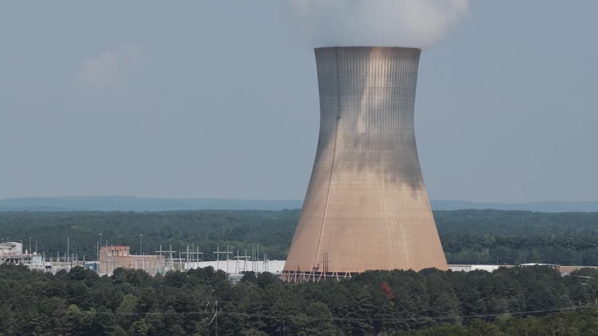 An aerial view slowly zooms out from a nuclear power plant's cooling tower as it releases steam into the sky