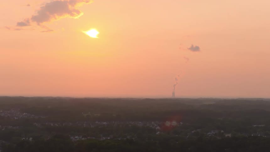 A hazy orange sunset over a forested landscape with a distant power plant smokestack emitting smoke