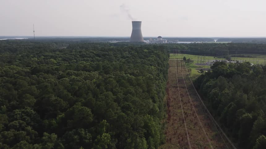 A slow aerial view moves over a dense forest and power lines towards a distant nuclear power plant