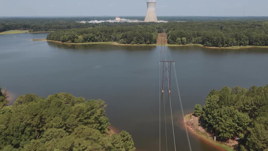 Aerial shot pulls back over a dense forest and lake to reveal power lines leading to a nuclear power plant