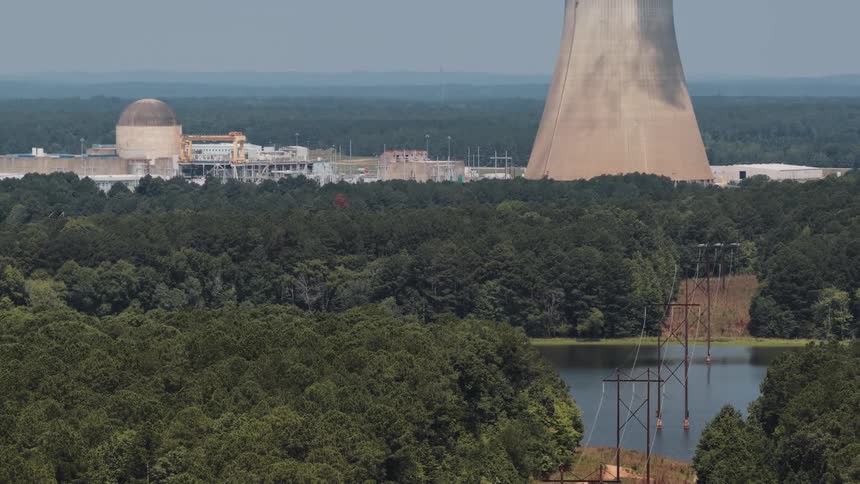 A slow aerial tilt-up reveals a nuclear power plant and its cooling tower behind a tranquil lake and forest