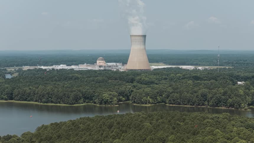 Wide static shot of a nuclear power plant's cooling tower emitting steam, surrounded by a lush green forest and a serene lake