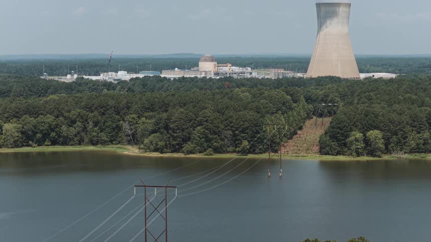 A slow aerial tilt reveals a nuclear power plant and its steaming cooling tower behind power lines crossing a lake
