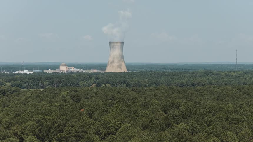 Static aerial view of a nuclear power plant cooling tower emitting steam above a vast green forest on a clear day