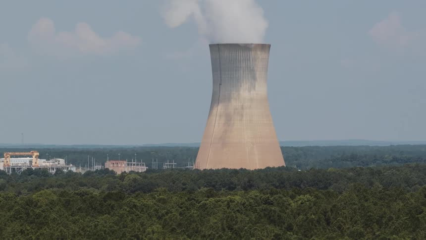 A slow aerial zoom out reveals a nuclear power plant's cooling tower emitting steam above a dense green forest