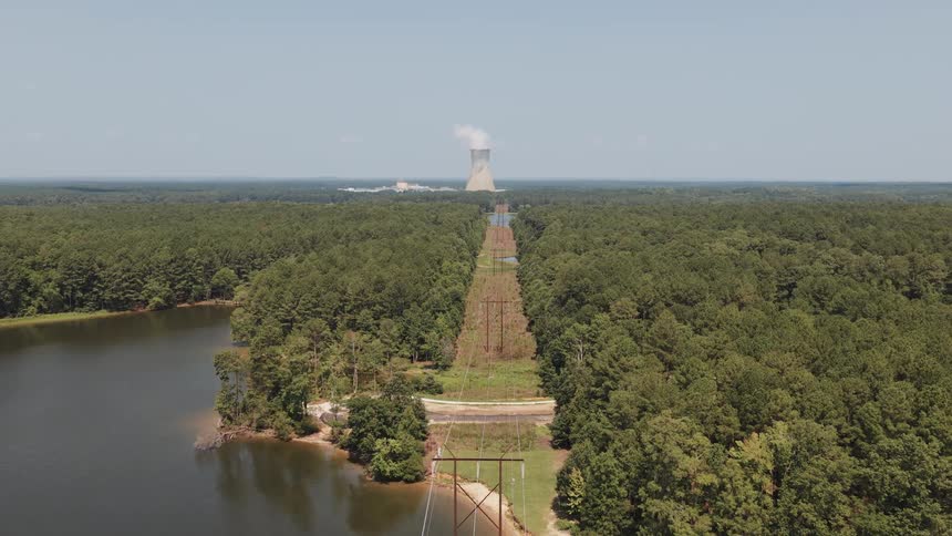 An aerial view shows power lines cutting a straight path through a dense forest and across a lake, leading to a nuclear power plant on the horizon