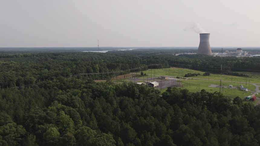 Wide aerial view of a nuclear power plant with a smoking cooling tower set in a vast rural forest landscape